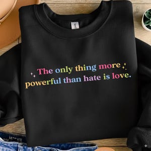 Puede incluir: Sudadera negra con la frase "The only thing more powerful than hate is love" en letras coloridas. El texto es rosa, azul, amarillo y verde. La sudadera está doblada sobre una superficie de madera.