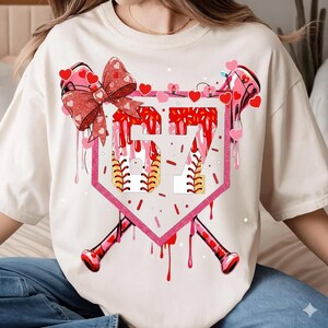 Puede incluir: Camiseta color crema con un diseño temático de béisbol con el número 57. El diseño incluye un plato de home de béisbol rosa, detalles rojos goteando, un lazo, corazones y bates de béisbol. Un gráfico festivo y deportivo.