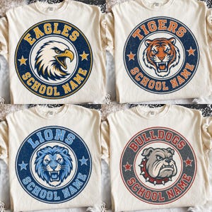 Puede incluir: Cuatro camisetas color hueso con estampados gráficos circulares. Cada camiseta muestra una cabeza de mascota (águila, tigre, león o bulldog) rodeada por el nombre del equipo y el texto SCHOOL NAME, con estrellas en un diseño vintage desgastado.
