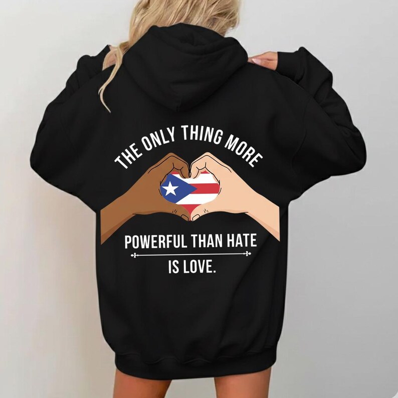 May include: Black hoodie with the text "THE ONLY THING MORE POWERFUL THAN HATE IS LOVE." The design features hands forming a heart around the Puerto Rican flag. The hoodie is a pullover style.