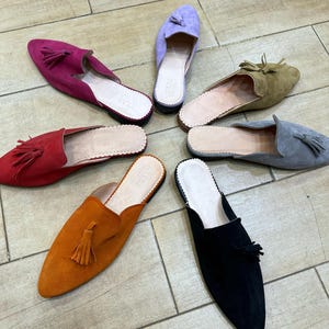 May include: A collection of suede slip-on shoes in various colors, including red, orange, black, gray, green, and purple. Each shoe features a tassel and a leather insole. The shoes are arranged in a circular pattern on a light brown tiled floor.