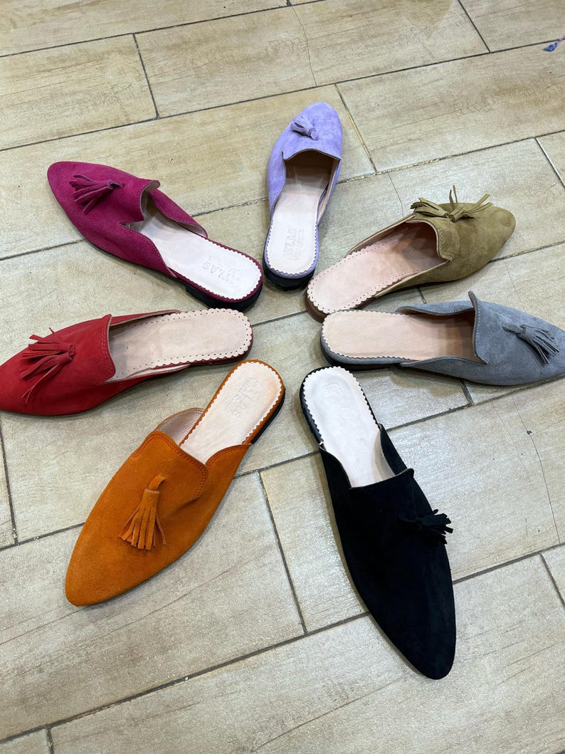 May include: A collection of suede slip-on shoes in various colors, including red, orange, black, gray, green, and purple. Each shoe features a tassel and a leather insole. The shoes are arranged in a circular pattern on a light brown tiled floor.
