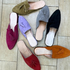 May include: An assortment of suede slip-on shoes in various colors, including olive green, purple, gray, black, red, and orange. Each shoe features a tassel and a rounded toe. The shoes are arranged on a tiled surface.