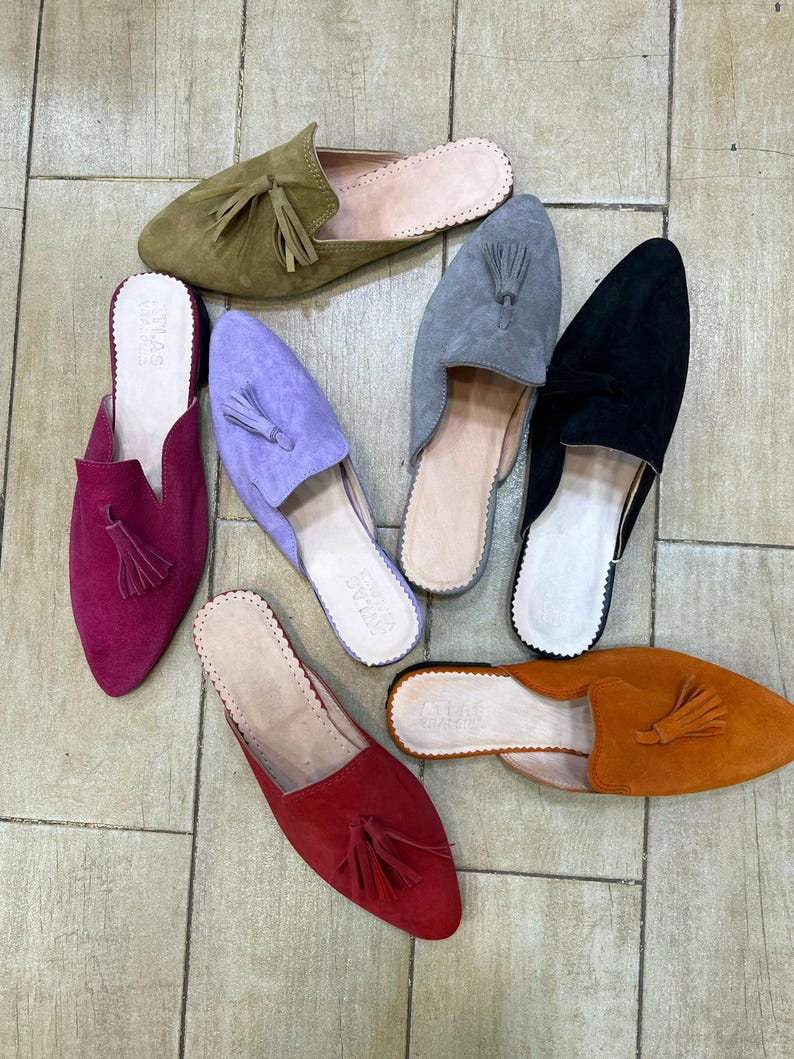 May include: An assortment of suede slip-on shoes in various colors, including olive green, purple, gray, black, red, and orange. Each shoe features a tassel and a rounded toe. The shoes are arranged on a tiled surface.
