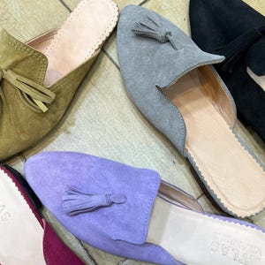 May include: A collection of suede slip-on shoes in various colors, including olive green, gray, black, and purple. Each shoe features a tassel detail and a scalloped edge around the opening. The shoes are arranged on a light-colored, tiled surface.