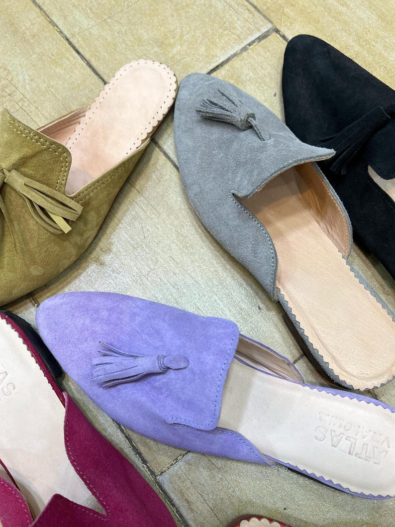May include: A collection of suede slip-on shoes in various colors, including olive green, gray, black, and purple. Each shoe features a tassel detail and a scalloped edge around the opening. The shoes are arranged on a light-colored, tiled surface.