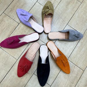 May include: A collection of suede slip-on shoes in various colors, including purple, green, gray, orange, red, black, and fuchsia. Each shoe features a tassel and a leather insole. The shoes are arranged in a circular pattern.