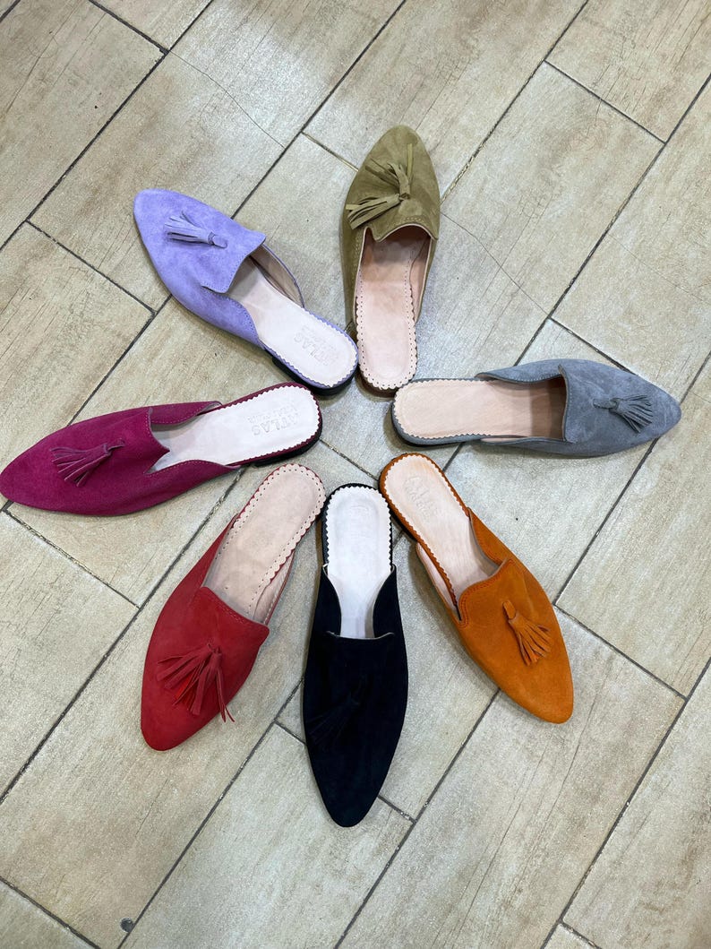 May include: A collection of suede slip-on shoes in various colors, including purple, green, gray, orange, red, black, and fuchsia. Each shoe features a tassel and a leather insole. The shoes are arranged in a circular pattern.