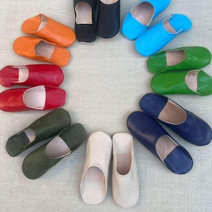 Puede incluir: Una colección de zapatillas de cuero coloridas en varios tonos, incluyendo naranja, rojo, negro, azul, verde y blanco. Las zapatillas están dispuestas en un patrón circular, mostrando sus diversos colores y diseños. Estas zapatillas de estilo marroquí son ideales para uso en interiores.