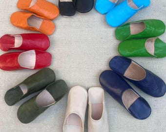 May include: A collection of colorful leather slippers in various shades, including orange, red, black, blue, green, and white. The slippers are arranged in a circular pattern, showcasing their diverse hues and designs. These Moroccan-style slippers are perfect for indoor wear.