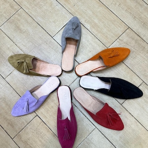 May include: A collection of colorful suede slip-on shoes arranged in a circular pattern. The shoes come in various colors, including olive green, lavender, orange, black, red, gray, and magenta. Each shoe features a pointed toe and a tassel.