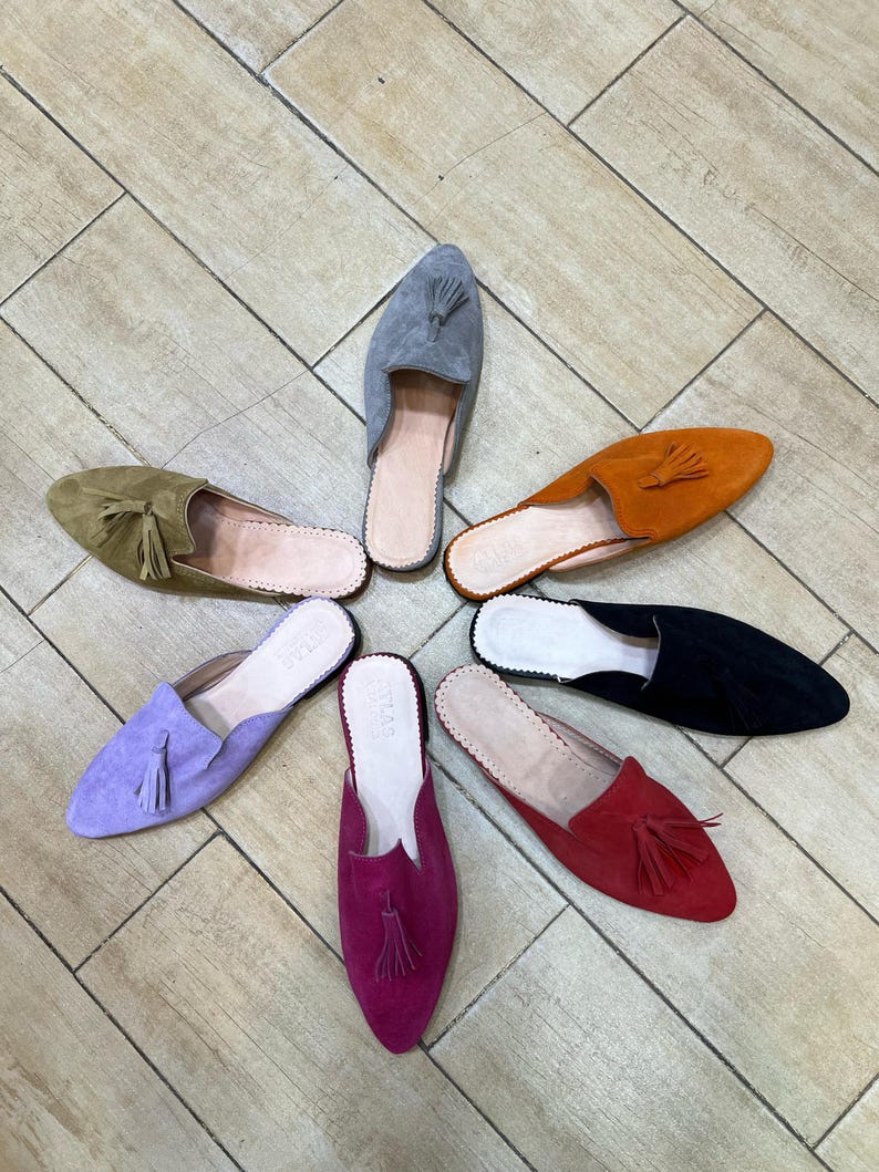 May include: A collection of colorful suede slip-on shoes arranged in a circular pattern. The shoes come in various colors, including olive green, lavender, orange, black, red, gray, and magenta. Each shoe features a pointed toe and a tassel.