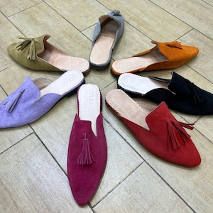 May include: A collection of colorful suede slip-on shoes with tassel accents. The shoes are in various colors, including olive green, lavender, orange, black, red, and fuchsia. The shoes are arranged in a circular pattern on a wooden floor.