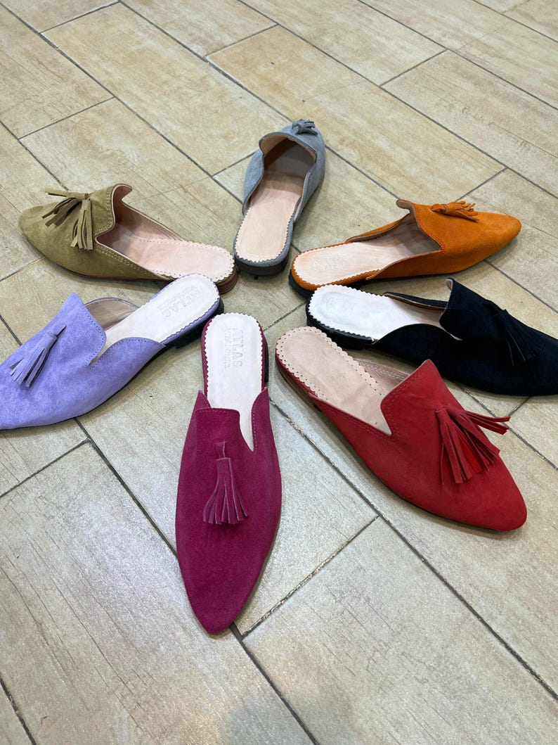 May include: A collection of colorful suede slip-on shoes with tassel accents. The shoes are in various colors, including olive green, lavender, orange, black, red, and fuchsia. The shoes are arranged in a circular pattern on a wooden floor.