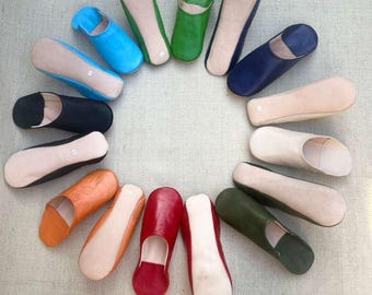Puede incluir: Una colección de zapatillas de cuero de colores dispuestas en un patrón circular. Las zapatillas vienen en varios colores, incluyendo azul, verde, negro, naranja, rojo y beige. Las suelas son de color beige claro.
