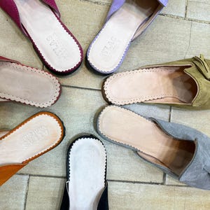 May include: Overhead shot of several colorful suede slippers. The slippers are in shades of pink, purple, orange, green, gray, and black. Each slipper has a scalloped edge and a leather insole. Some have tassels.