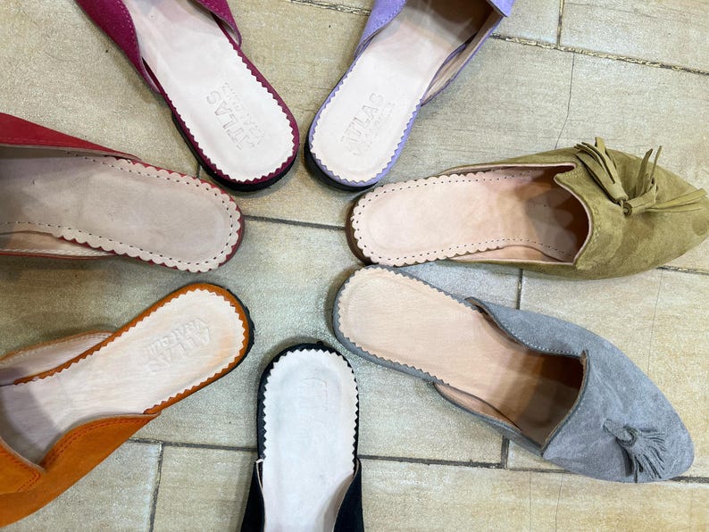 May include: Overhead shot of several colorful suede slippers. The slippers are in shades of pink, purple, orange, green, gray, and black. Each slipper has a scalloped edge and a leather insole. Some have tassels.