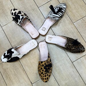 May include: Five pairs of slip-on shoes with animal print designs. The patterns include cowhide, zebra, and leopard prints. Each shoe has a black tassel and a light pink insole. The shoes are arranged on a light brown wooden floor.