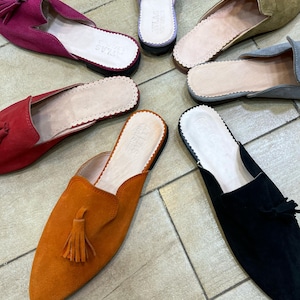 May include: A collection of colorful suede slip-on shoes with pointed toes and tassels. The shoes are in various colors, including fuchsia, red, orange, black, gray, olive green, and lavender. The insoles are light-colored with a scalloped edge.