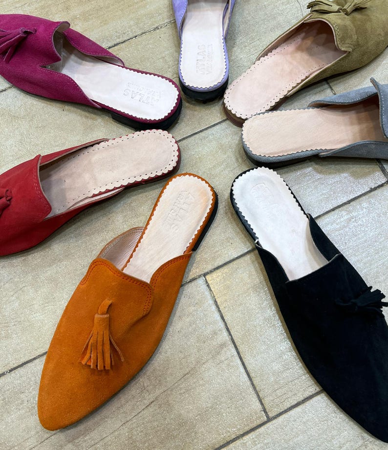 May include: A collection of colorful suede slip-on shoes with pointed toes and tassels. The shoes are in various colors, including fuchsia, red, orange, black, gray, olive green, and lavender. The insoles are light-colored with a scalloped edge.