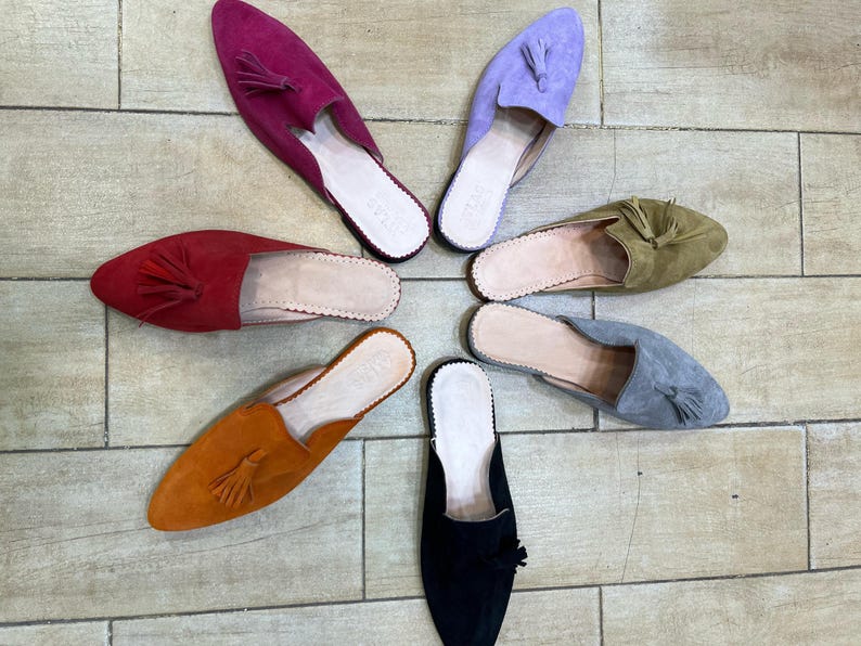 May include: A collection of colorful suede slip-on shoes arranged in a circular pattern. The shoes come in various colors, including red, purple, orange, green, gray, and black. Each shoe features a pointed toe and a tassel detail.