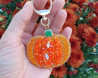 May include: A pumpkin-shaped keychain featuring vibrant orange beads, faceted crystals, and a small green beaded stem. The ornament is attached to a polished silver-tone metal swivel clasp and ring, showcasing intricate beadwork and reflective textures.