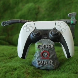 May include: A white PlayStation 5 DualSense controller is resting on a grey stone base with the words "God of War" printed on it. The controller has a black and white colour scheme and features a black and white axe.