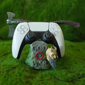May include: A white PlayStation 5 controller sits on a grey rock stand with the words "God of War" printed on it. The controller is connected to a black and silver axe.