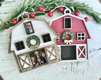 May include: Two wooden barn-shaped ornaments with family photos. One barn is white with a green wreath and a light brown bow, the other is red with a green wreath and a light brown bow. Both have black windows and are decorated with a sprig of greenery and red berries.