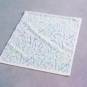 May include: White square textile with a repeating pattern of blue and teal abstract shapes. The fabric is folded, and the edges are finished with a white border. The textile is displayed on a gradient grey and blue surface.