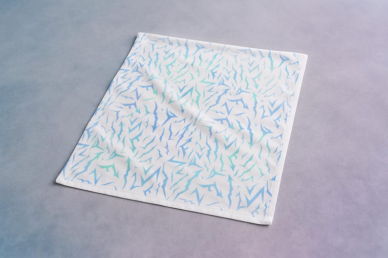 May include: White square textile with a repeating pattern of blue and teal abstract shapes. The fabric is folded, and the edges are finished with a white border. The textile is displayed on a gradient grey and blue surface.