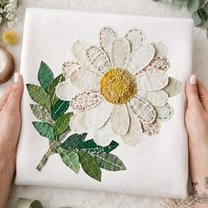 May include: A white fabric square featuring a large daisy applique. The daisy has off-white petals with varied textures and a yellow center. Green leaves and stem are on the left. The piece is held by hands.