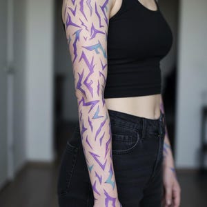 May include: A full arm tattoo with a geometric design in purple and teal. The tattoo features abstract shapes, creating a modern look. The person is wearing a black vest top and black jeans. The tattoo is a striking piece of body art.