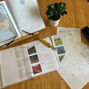 May include: An open three-ring binder displays a map of Western Africa and a geography worksheet with images of a hill, delta, and desert. Nearby are loose worksheets, three yellow pencils, a black hole punch, and a small potted green plant.