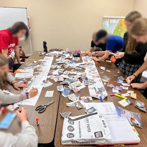 Può includere: Un gruppo di persone che lavorano insieme a un grande tavolo marrone coperto di carta, immagini e forniture. Sono visibili forbici, stick di colla e carta da copia. L'attività sembra essere un progetto o una presentazione collaborativa.