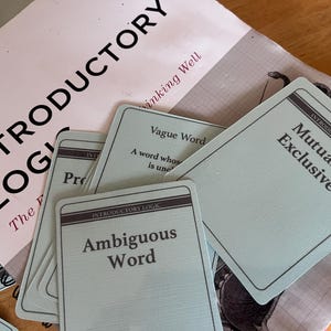 May include: A selection of logic flashcards and a book titled "Introductory Logic". The cards are pale green with black text, displaying terms such as "Ambiguous Word" and "Mutually Exclusive". The book cover is pink with the title in large black letters.