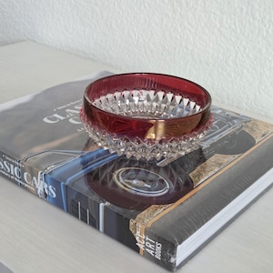 May include: A round glass bowl featuring a deep red rim and a clear, faceted diamond-cut pattern on the lower exterior. The bowl has a smooth, circular opening and a textured base, showcasing a contrast between the colored glass and clear crystal.