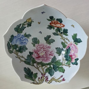 May include: A white ceramic bowl with a scalloped, petal-like edge. The interior features a hand-painted design of blooming peonies in shades of blue, pink, and orange, with green leaves, brown branches, and small butterflies scattered throughout.
