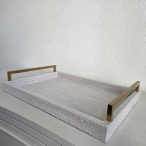 May include: A rectangular serving tray featuring a light gray, textured fabric base and two metallic gold-toned handles attached to the shorter sides. The tray has a clean, structured design with a flat bottom and raised edges for containment.