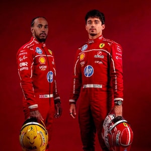 May include: Two individuals in red racing suits, each holding a helmet. The suits feature sponsor logos and white stripes. One helmet is yellow with a black design, the other is red with white accents. The background is a solid red.