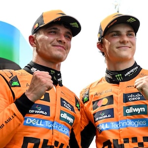 May include: Two people wearing orange racing suits and caps, with various sponsor logos. The suits have black and white checkered patterns. The individuals are smiling and clenching their fists. The background is a bright, sunny day.