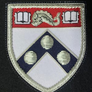 Könnte beinhalten: Ein silber gesticktes Wappen mit weißem Hintergrund und einem blauen Chevron mit drei silbernen Kreisen. Der obere Teil des Wappens zeigt ein rotes Banner mit zwei offenen Büchern und einem silbernen stilisierten Fisch.