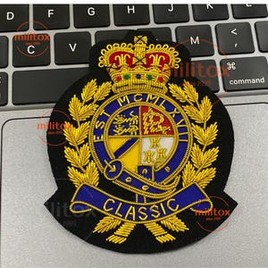以下が含まれることがあります： 金色の王冠、月桂樹の葉、中央のエンブレムが付いた黒色の刺繍パッチ。エンブレムは、青と黄色のデザインで、「ST MCMLXVII」と「CLASSIC」の文字が青いバナーに書かれています。パッチはキーボードの上にあります。