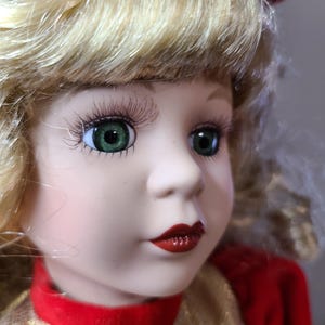 May include: A porcelain doll with blonde hair and green eyes. The doll has long eyelashes, red lips, and is wearing a red and gold dress. A red bow is in the doll's hair.