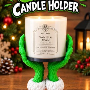 May include: A green candle holder shaped like two hands with white fur cuffs and red pleated sleeves, supporting a white cylindrical candle jar labelled Vanilla Bean with 14.5 oz and 411 g text. The text Grinch Candle Holder appears above.