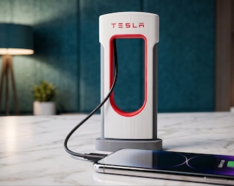 Pode incluir: Uma estação de carregamento de mesa estilo Tesla Supercharger branca e cinza com o logotipo vermelho da Tesla no topo. A unidade possui uma estrutura central aberta com detalhes internos vermelhos e um cabo preto conectado a um smartphone.