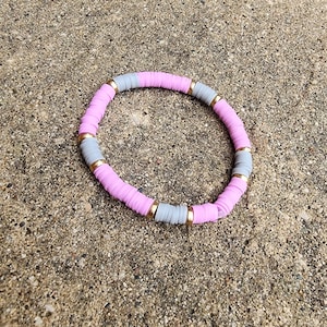 May include: A pink and gray beaded bracelet with gold accents. The bracelet is made of round beads and is strung on a stretchy cord.