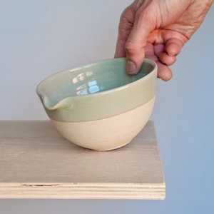 May include: A handcrafted ceramic bowl with a pale green glazed upper section and a natural, unglazed base. The bowl features a spout and is held by a hand, highlighting its shape and design. The bowl is placed on a wooden surface.