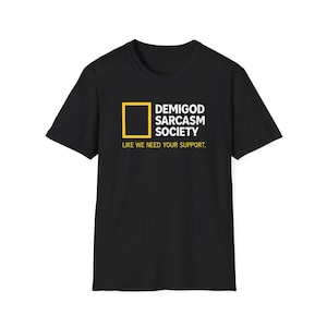 Peut inclure: T-shirt noir avec l'inscription blanche "DEMIGOD SARCASTIC SOCIETY" et un carré jaune. La phrase "LIKE WE NEED YOUR SUPPORT" est imprimée en dessous.
