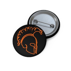May include: Two round black button pins. One pin features an orange outline of a Spartan helmet. The other pin is silver with a pin back. The helmet design is a simple line drawing.
